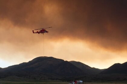 A firefighting helicopter flying near heavy smoke columns rising from a western National Forest wildfire