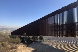 Tall steel bollard border barrier fence stretching across desert terrain along the southwest border